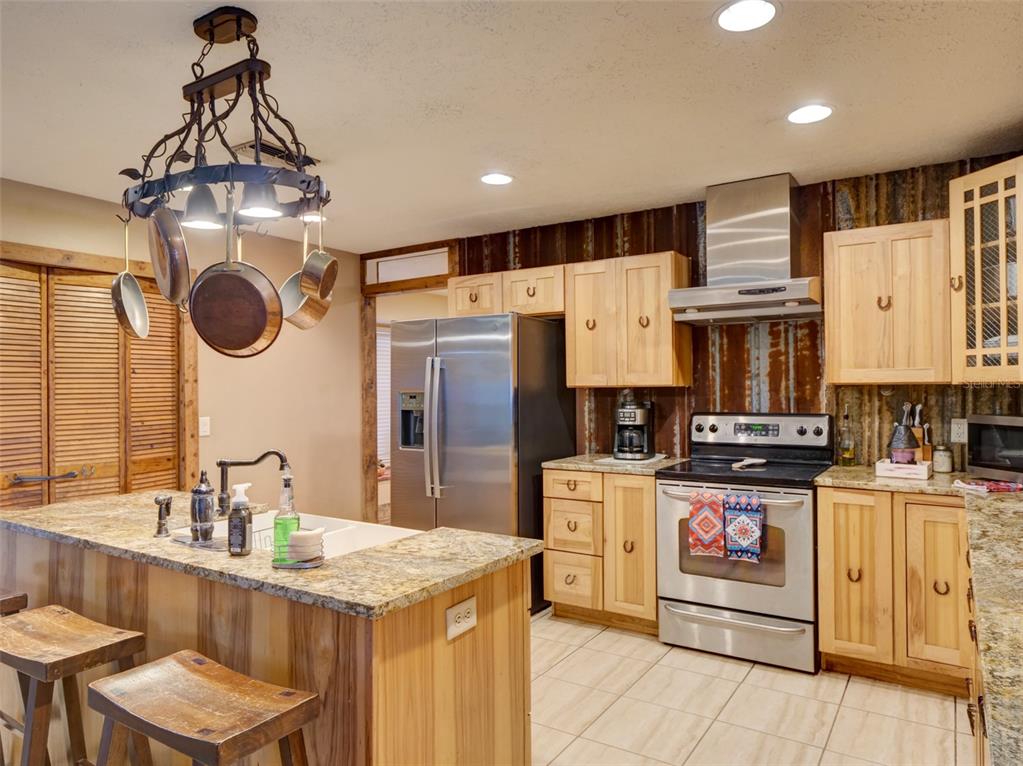 A rustic kitchen features wood cabinets, granite countertops, stainless steel appliances, and a hanging pot rack — 1765 E CR 462, WILDWOOD, FL