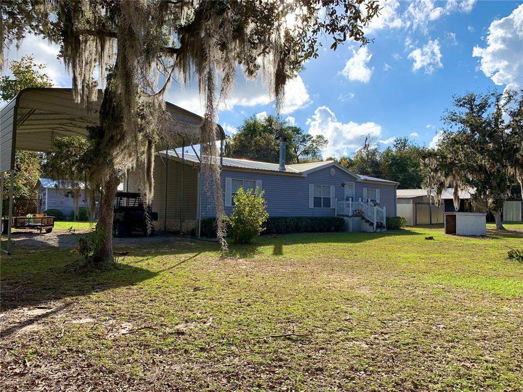 A single-story manufactured home with a metal carport and Spanish moss-covered trees in the yard — 17630 NE 246TH PLACE, FORT MC COY, FL