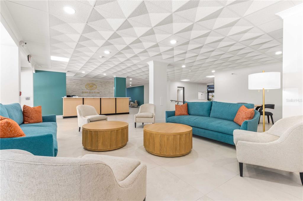 A modern lobby features teal sofas, light-colored armchairs, round wooden coffee tables, and a textured ceiling — 1380 GULF BOULEVARD UNIT 805, CLEARWATER, FL