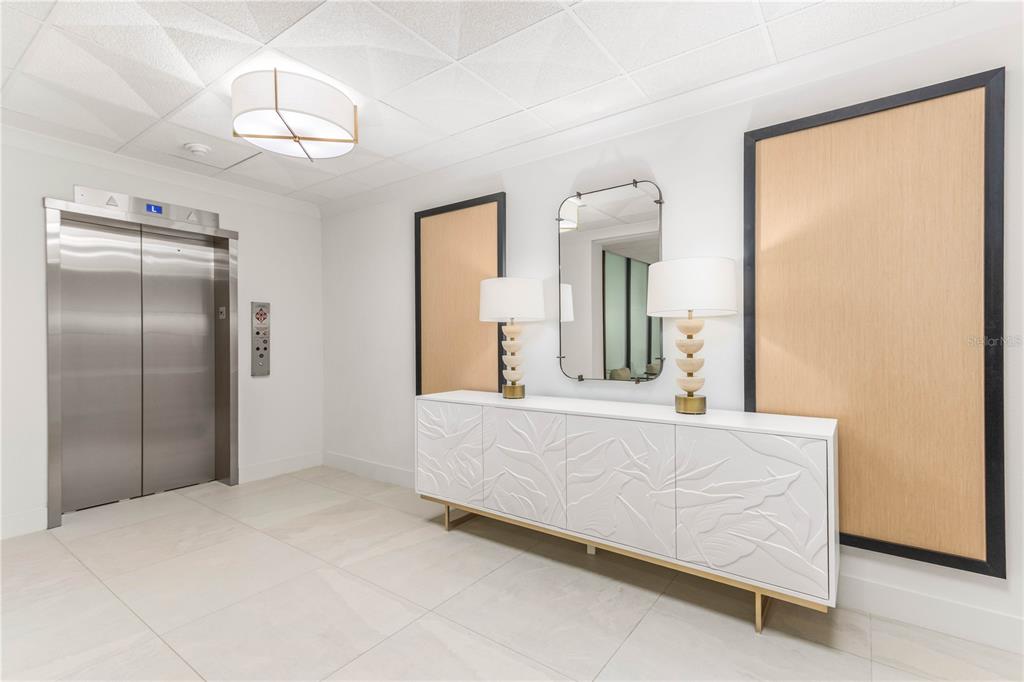 A modern elevator lobby features a white textured sideboard, two lamps, a mirror, and panels — 1380 GULF BOULEVARD UNIT 805, CLEARWATER, FL