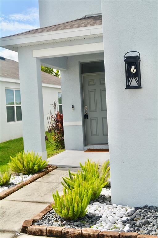 A light-colored house exterior features a covered entryway, a grey front door, and landscaped garden beds — 34110 JASPER STONE DRIVE, WESLEY CHAPEL, FL