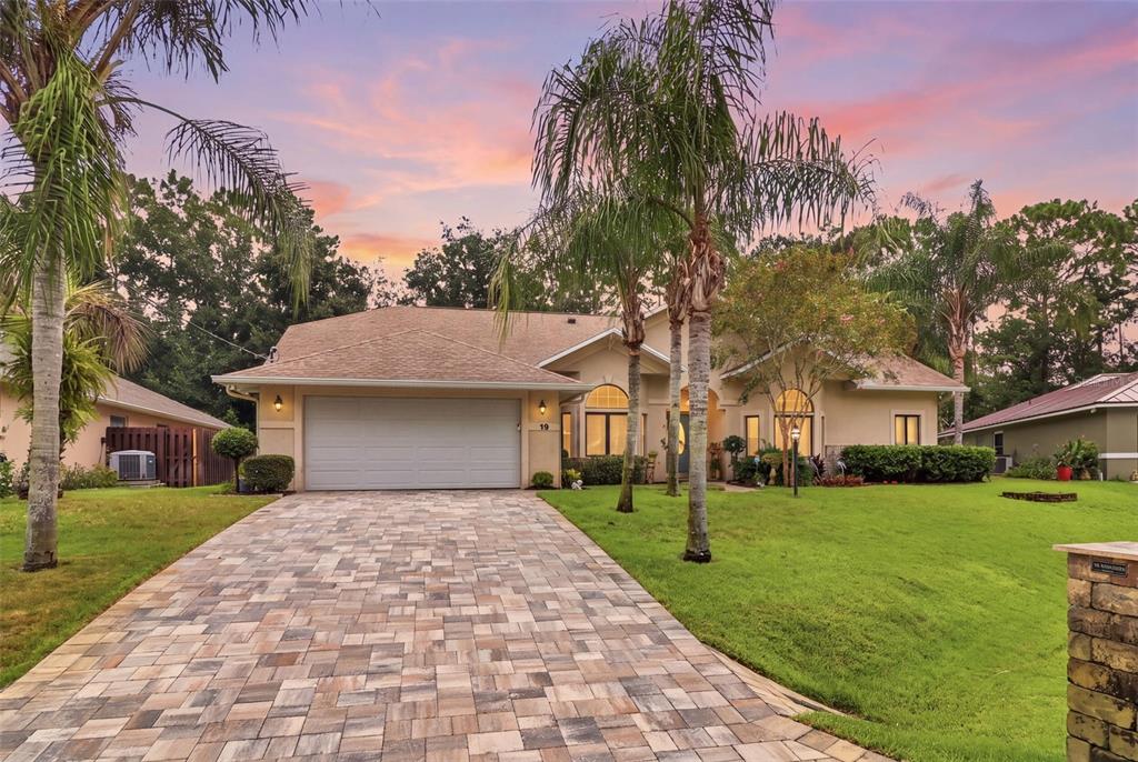 A single-story house with a paved driveway, palm trees, and a two-car garage at sunset — 19 WOODGUILD PLACE, PALM COAST, FL