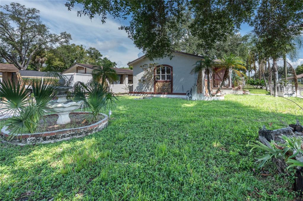 A grassy yard features a stone fountain, palm plants, and a single-story house with siding — 5104 N 34TH STREET, TAMPA, FL