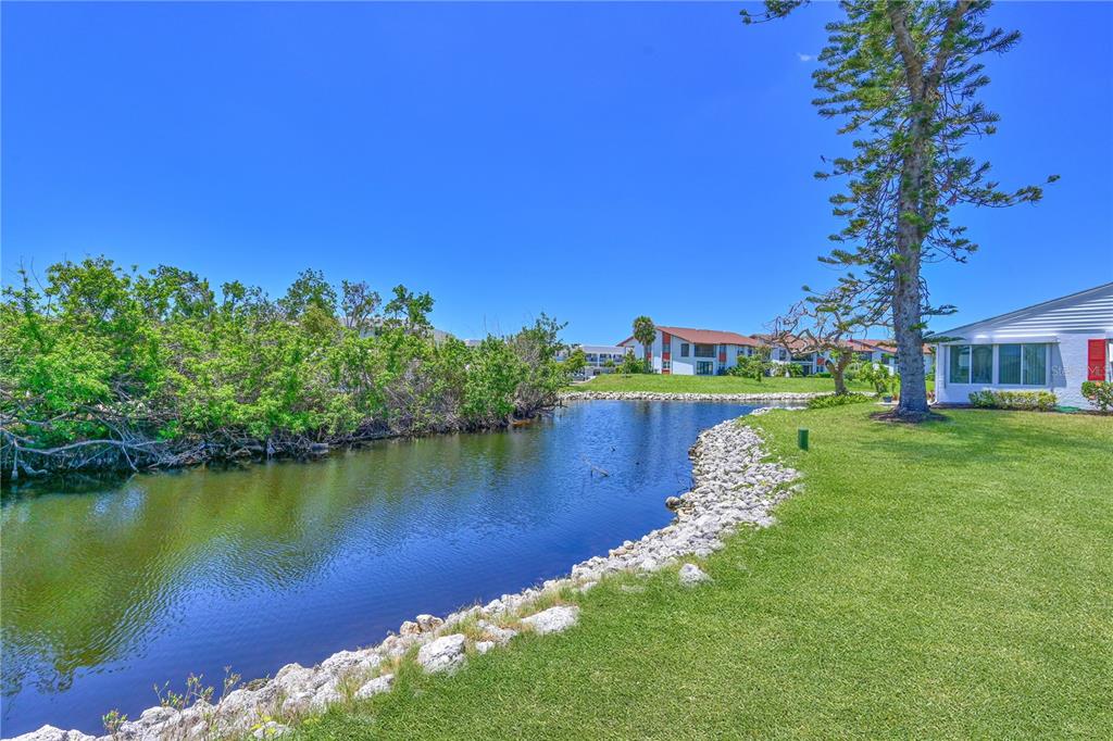 A grassy lawn overlooks a small pond with a rocky edge and distant apartment buildings — 4726 POTOMAC CIRCLE, BRADENTON, FL