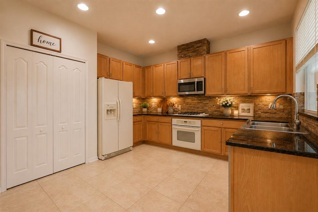 A kitchen features light wood cabinets, dark granite countertops, a stone backsplash, and white appliances — 4716 MAPLETREE LOOP, WESLEY CHAPEL, FL