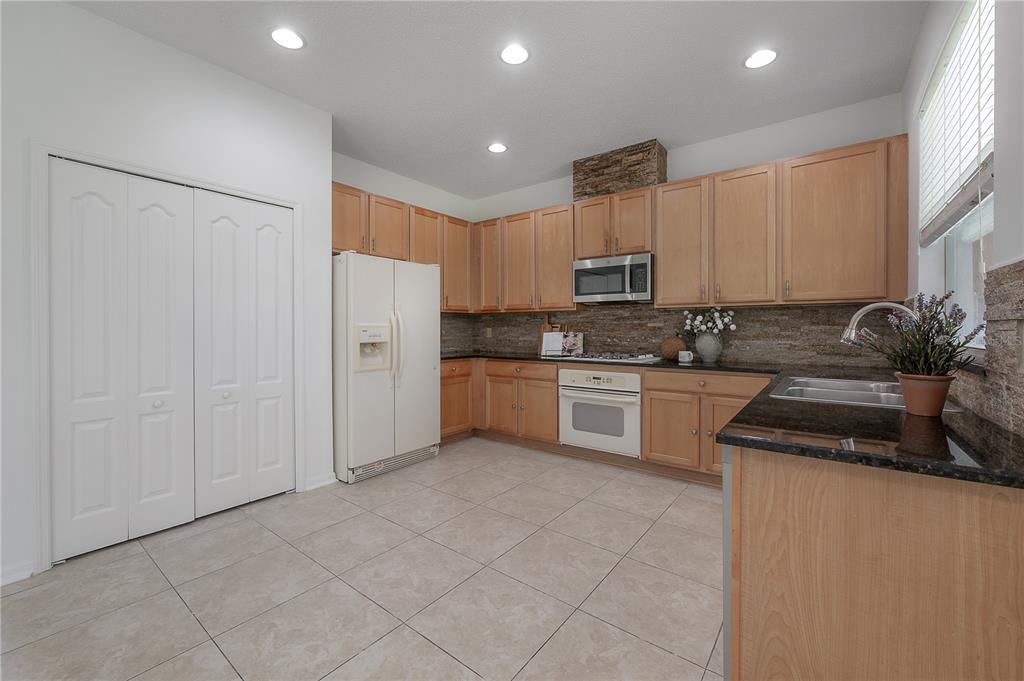 A kitchen with light wood cabinets, dark countertops, and tile flooring opens to a living area — 4716 MAPLETREE LOOP, WESLEY CHAPEL, FL