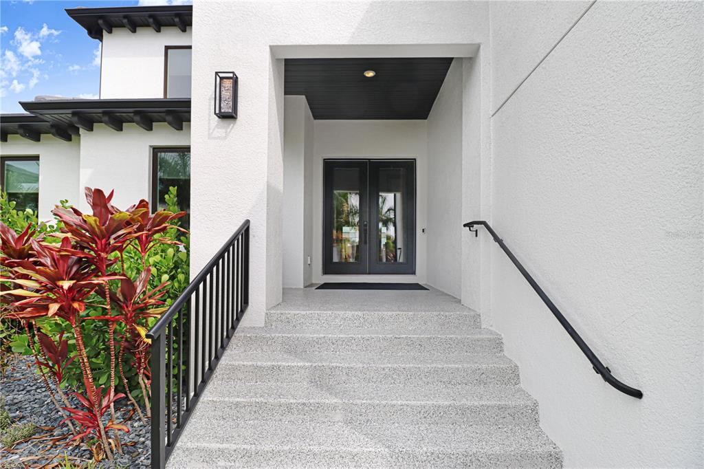 A modern white house entrance features concrete stairs, black railings, and double glass doors — 6104 LAGOMAR LANE, APOLLO BEACH, FL