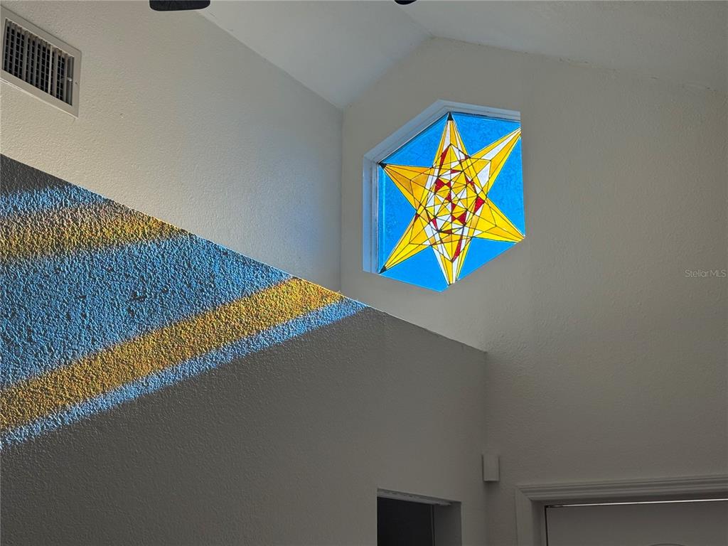 A high-ceilinged room features a hexagonal stained glass window casting colorful light onto the wall — 7309 N HUNTLEY AVENUE, TAMPA, FL
