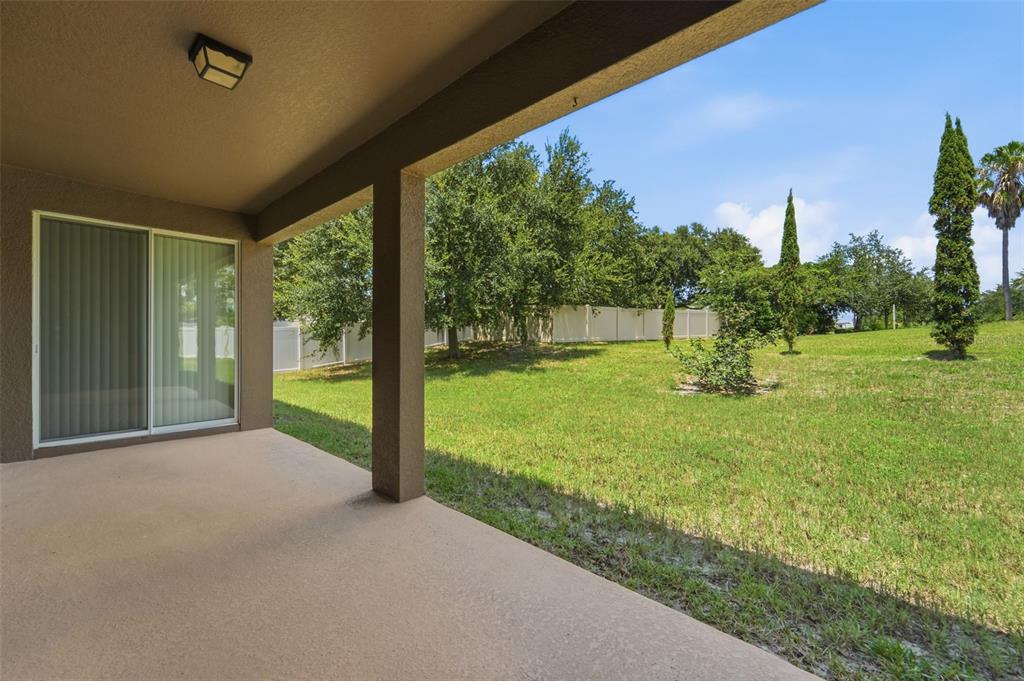 A covered concrete patio overlooks a grassy backyard with trees and a white vinyl fence — 1451 DAYSTAR LANE, DELTONA, FL