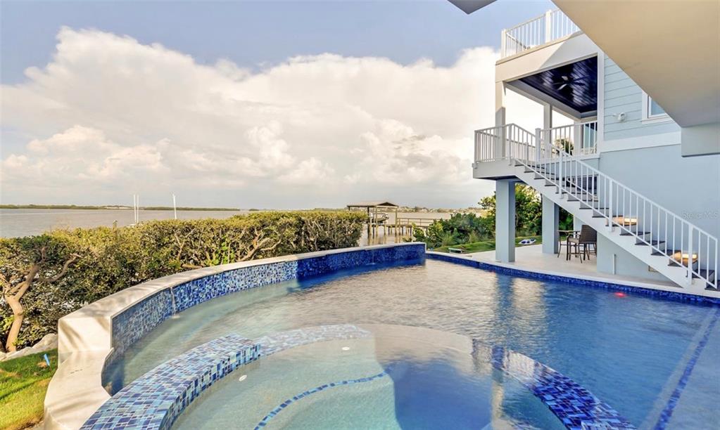 A waterfront infinity pool and hot tub sit beside a multi-story house with exterior stairs — 2516 AVENUE A, BRADENTON BEACH, FL