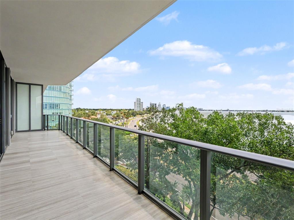 A modern balcony features light-colored tile flooring, a glass railing, and a view of trees and water — 3101 W BAYSHORE STREET UNIT 502, TAMPA, FL