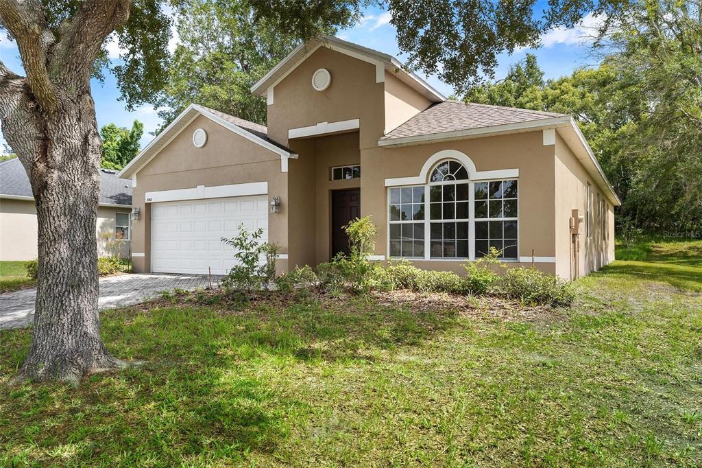 A tan, single-story suburban house with a two-car garage and a large front tree — 3360 CREEK RUN LANE, EUSTIS, FL