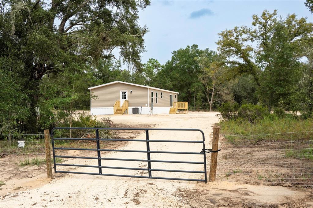 A tan mobile home sits behind a metal gate on a sandy, wooded lot — 11091 NE 80 STREET, BRONSON, FL
