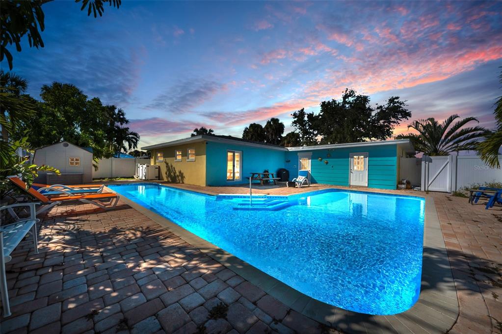A backyard features a swimming pool, a blue house, and a paved patio at sunset — 424 88TH AVENUE, ST PETE BEACH, FL