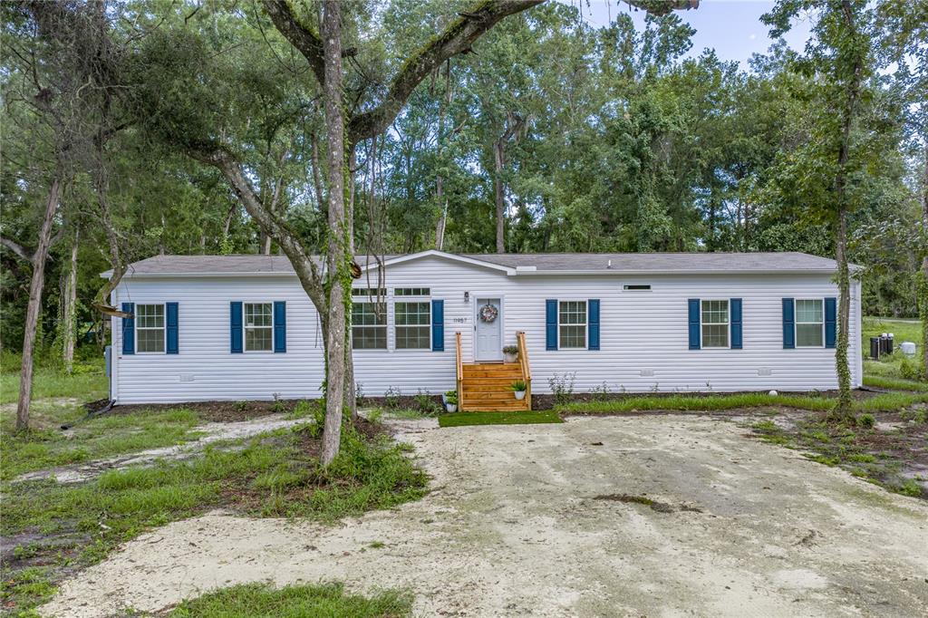 A single-story manufactured home with white siding and blue shutters sits in a wooded lot — 11957 SW 35TH STREET, WEBSTER, FL