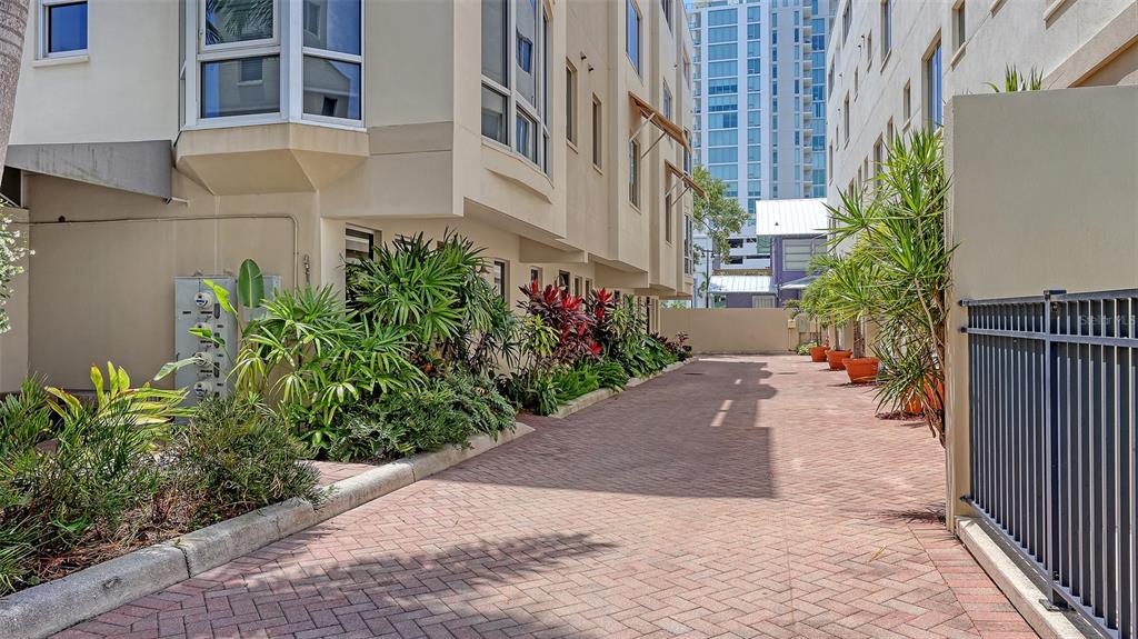 A brick-paved alleyway runs between two multi-story buildings with tropical landscaping and potted plants — 235 COCOANUT AVENUE UNIT 128E, SARASOTA, FL