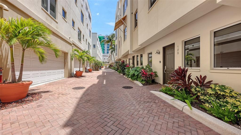 A paved alleyway between two modern multi-story buildings with garage doors and landscaped planters — 235 COCOANUT AVENUE UNIT 128E, SARASOTA, FL
