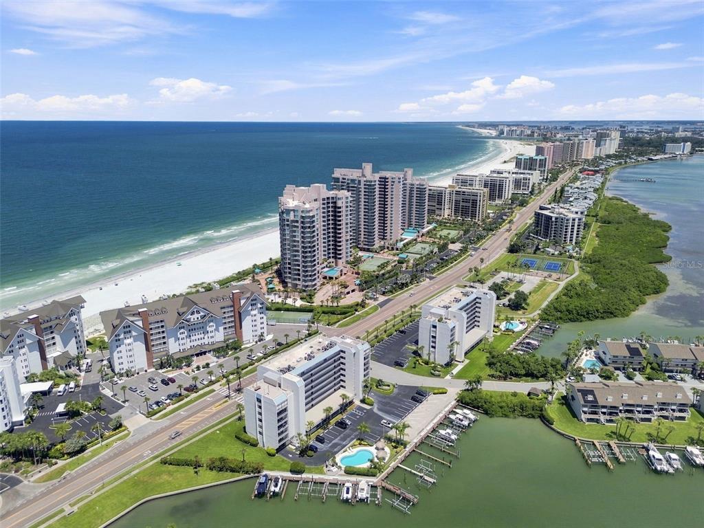An aerial view shows high-rise condominiums along a beach and a coastal waterway — 1582 GULF BOULEVARD UNIT 1106, CLEARWATER, FL