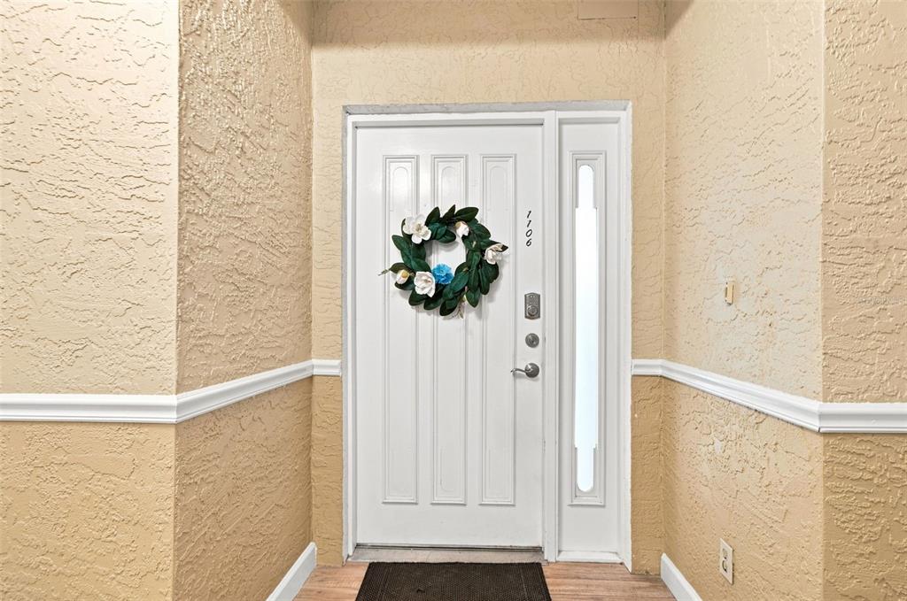 A white front door with a floral wreath sits between beige walls with white trim — 1582 GULF BOULEVARD UNIT 1106, CLEARWATER, FL
