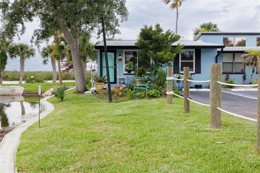 A light blue cottage with a metal roof sits beside a grassy lawn and water — 100 POINCIANA COVE COURT UNIT 100, NEW SMYRNA BEACH, FL