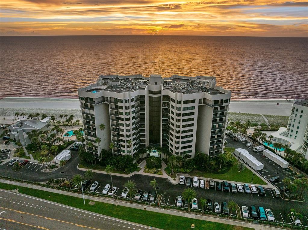 An aerial view shows a multi-story beachfront condominium building with a parking lot at sunset — 1600 GULF BOULEVARD UNIT 617, CLEARWATER BEACH, FL