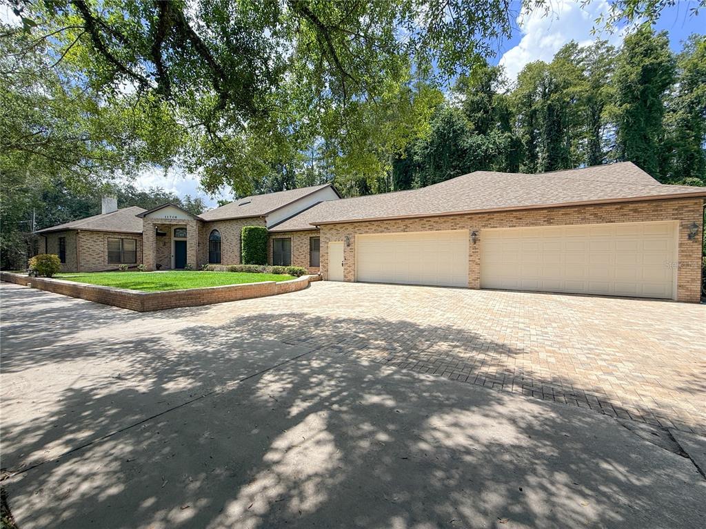 A brick house with a three-car garage and a paved driveway under large shade trees — 11708 ORANGE GROVE DRIVE, TAMPA, FL