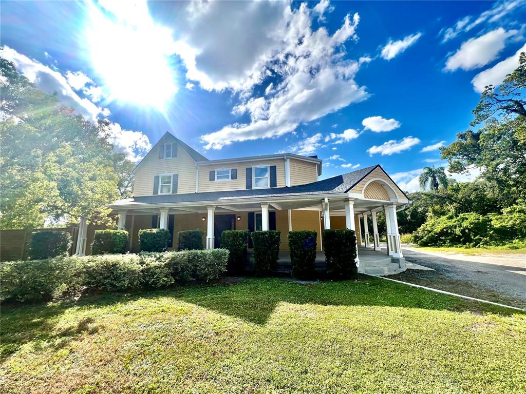 A two-story yellow house features a wrap-around porch with white columns and a gravel driveway — 1109 BELCHER ROAD, PALM HARBOR, FL