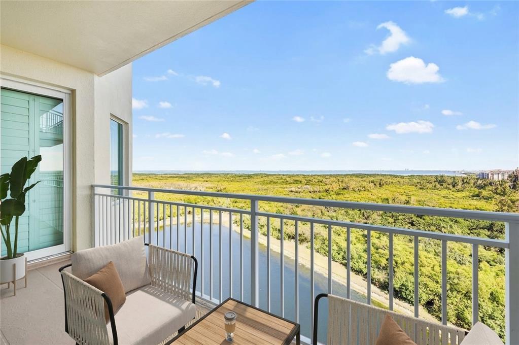 A balcony with outdoor seating overlooks a pond, dense green vegetation, and the distant ocean — 10851 MANGROVE CAY LANE NE UNIT 1112, ST PETERSBURG, FL