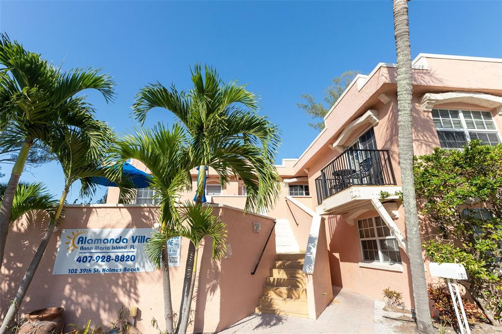 A peach-colored multi-story building features a staircase leading to an upper balcony with palm trees — 102 39TH STREET UNIT 2, HOLMES BEACH, FL