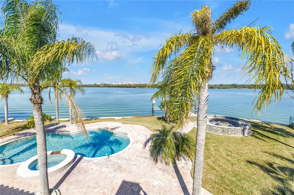 A backyard features a swimming pool, palm trees, and a fire pit overlooking a lake — 583 CRYSTAL DRIVE, MADEIRA BEACH, FL