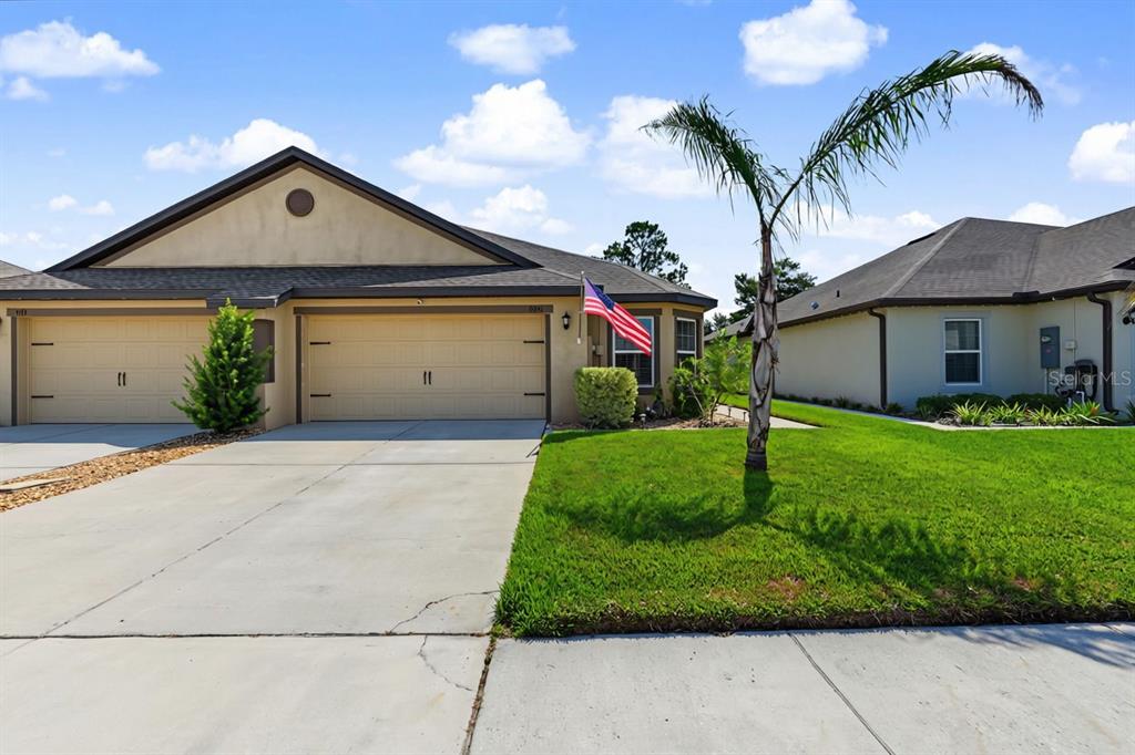 A tan single-story house with a two-car garage, a palm tree, and an American flag — 8951 SOUTHERN CHARM CIRCLE, BROOKSVILLE, FL