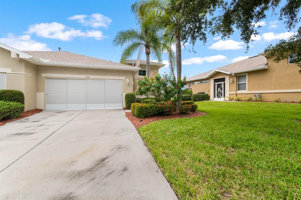 A concrete driveway leads to a two-car garage next to a tan house with landscaping — 1160 JAMESON GREENS DRIVE, SUN CITY CENTER, FL