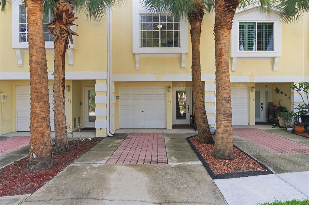 A row of three yellow townhouses with garages, palm trees, and brick-paved driveways — 117 BRENT CIRCLE, OLDSMAR, FL
