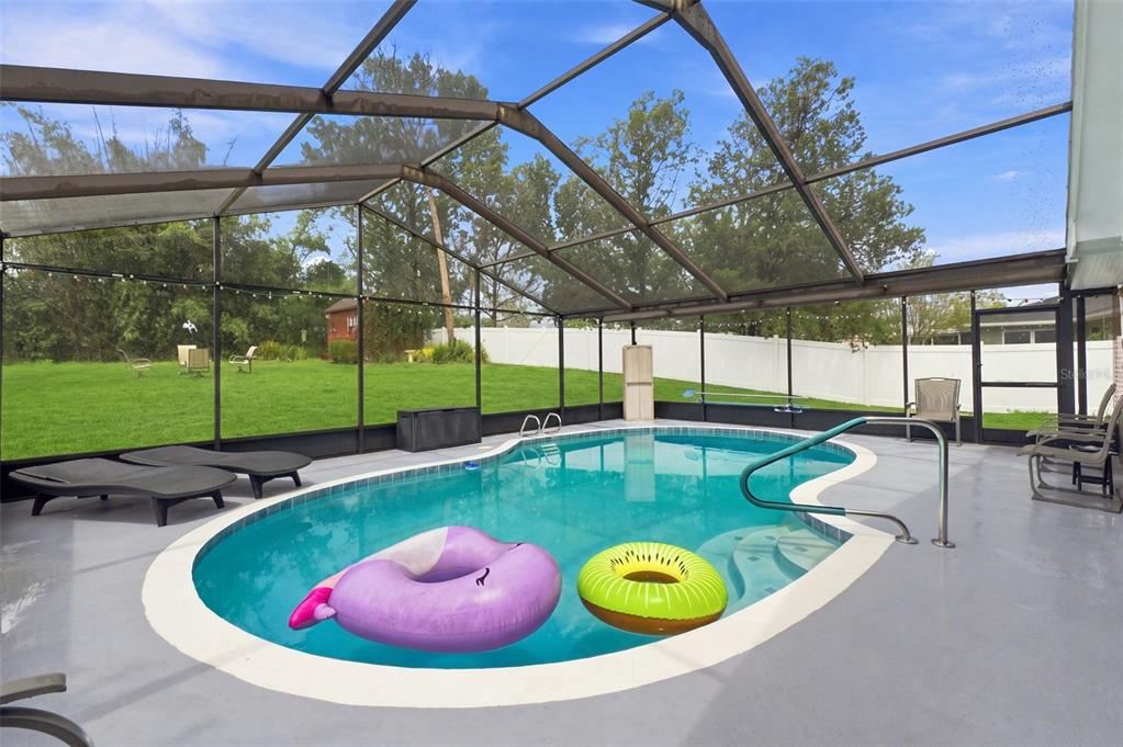 A screened-in swimming pool with two inflatable floats sits on a patio overlooking a backyard — 12411 BROOKSIDE STREET, SPRING HILL, FL