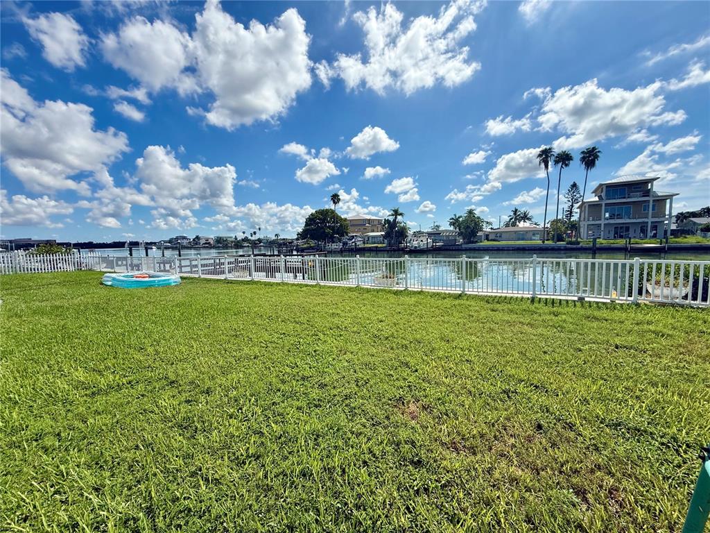 A grassy backyard features a white fence overlooking a canal with houses and palm trees — 722 PRUITT DRIVE, MADEIRA BEACH, FL
