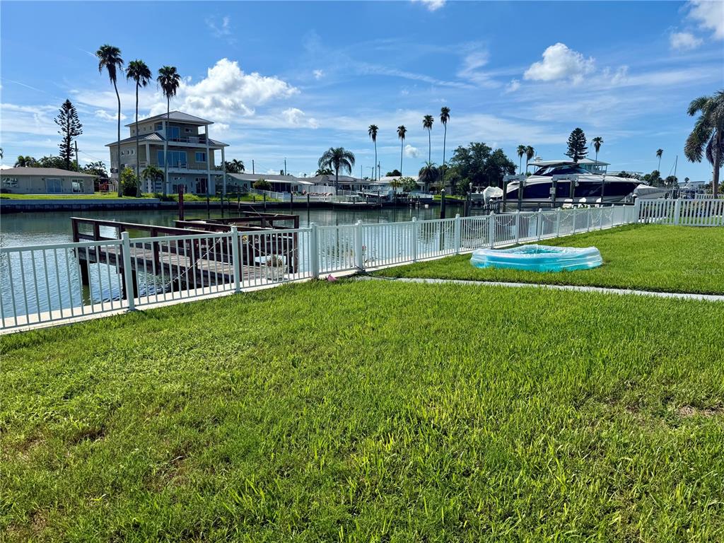 A grassy backyard features a white fence, a wooden dock, and a canal view — 722 PRUITT DRIVE, MADEIRA BEACH, FL