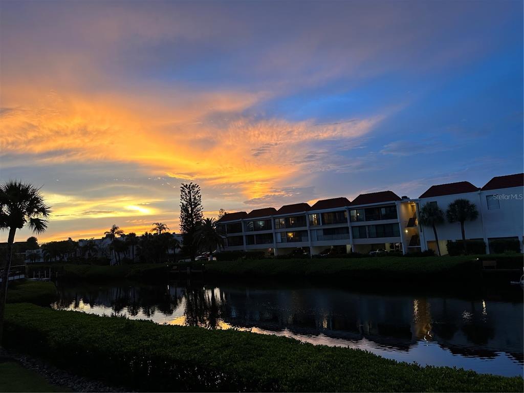 A multi-story apartment building sits beside a pond under a vibrant sunset sky — 3705 E BAY DRIVE UNIT 103, BRADENTON BEACH, FL