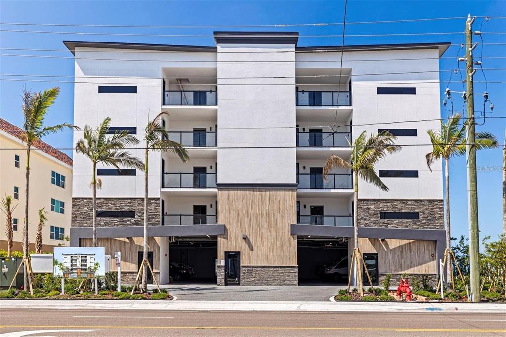 A modern four-story apartment building with a ground-level parking garage and palm trees in front — 14048 GULF BOULEVARD UNIT 402, MADEIRA BEACH, FL