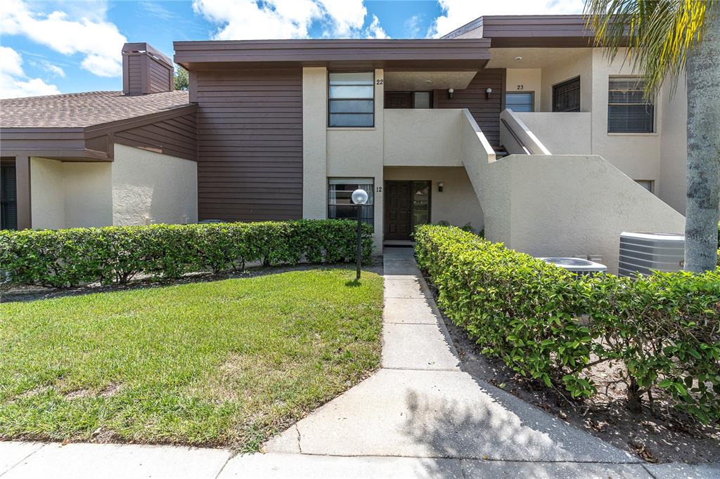 A two-story apartment building exterior features a concrete walkway, manicured hedges, and unit numbers — 2866 COUNTRYBROOK DRIVE UNIT 12, PALM HARBOR, FL