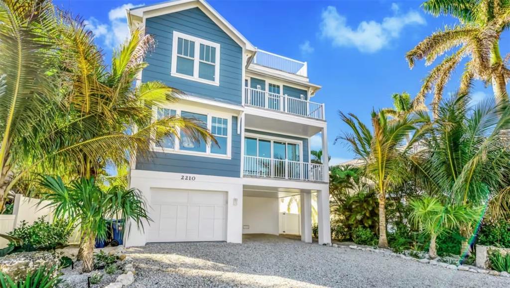 A three-story blue house with a white garage and balconies stands among palm trees — 2210 AVENUE A, BRADENTON BEACH, FL