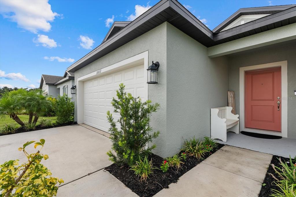 A light-colored stucco house exterior features a white garage door, a coral front door, and landscaping — 10032 LAST LIGHT GLEN, PARRISH, FL