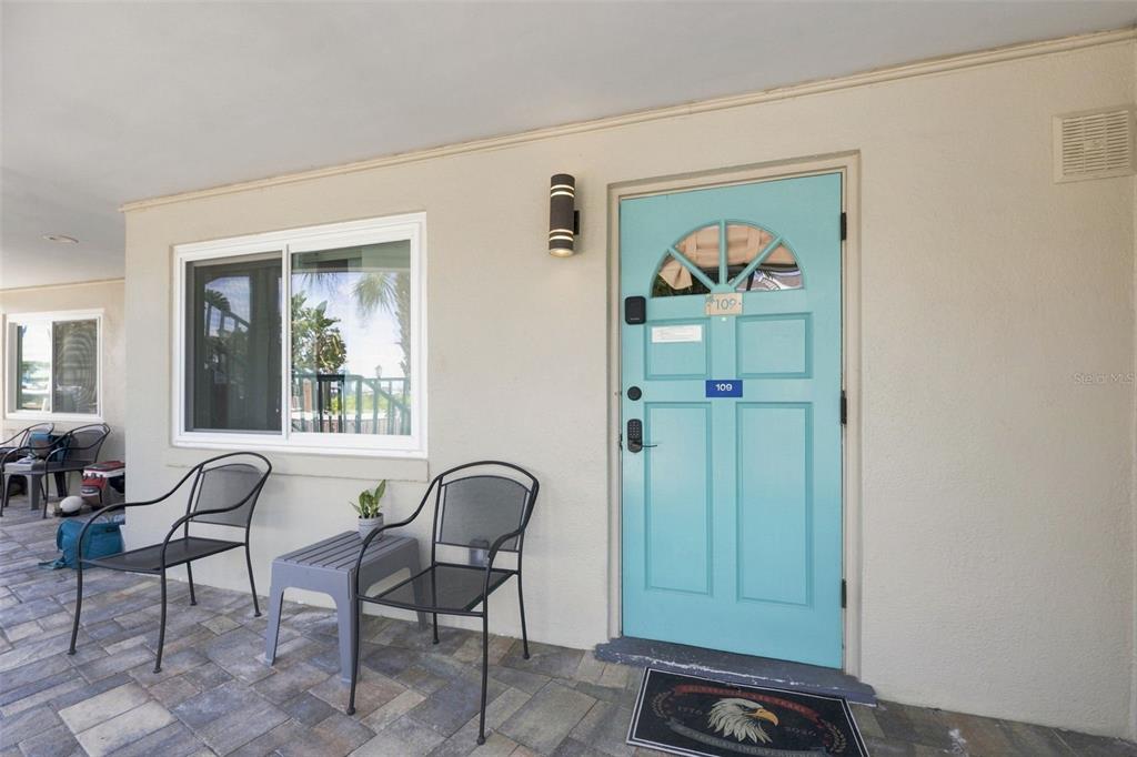 A light blue front door with a small window sits beside a window and chairs — 673 BAY ESPLANADE UNIT 109, CLEARWATER, FL