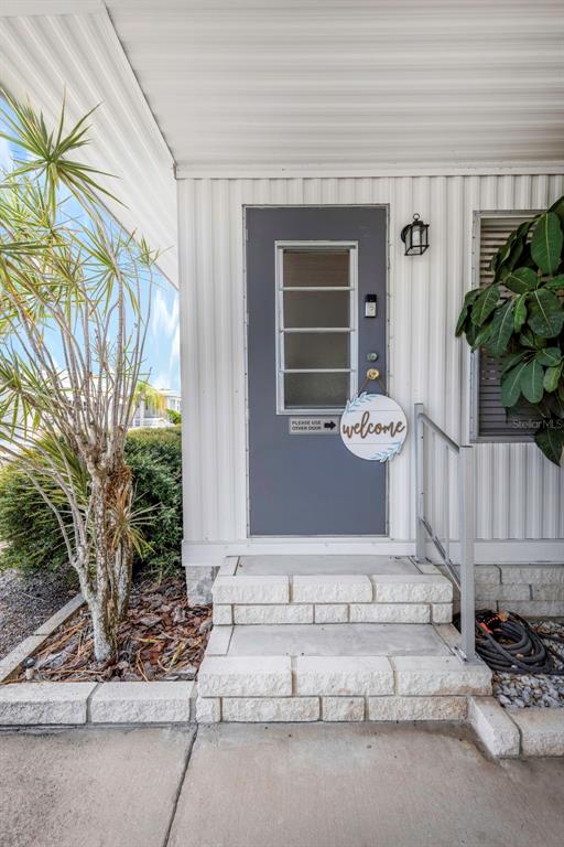 A gray front door with a welcome sign sits above concrete steps under a porch — 3301 US ALT 19 N UNIT 253, DUNEDIN, FL