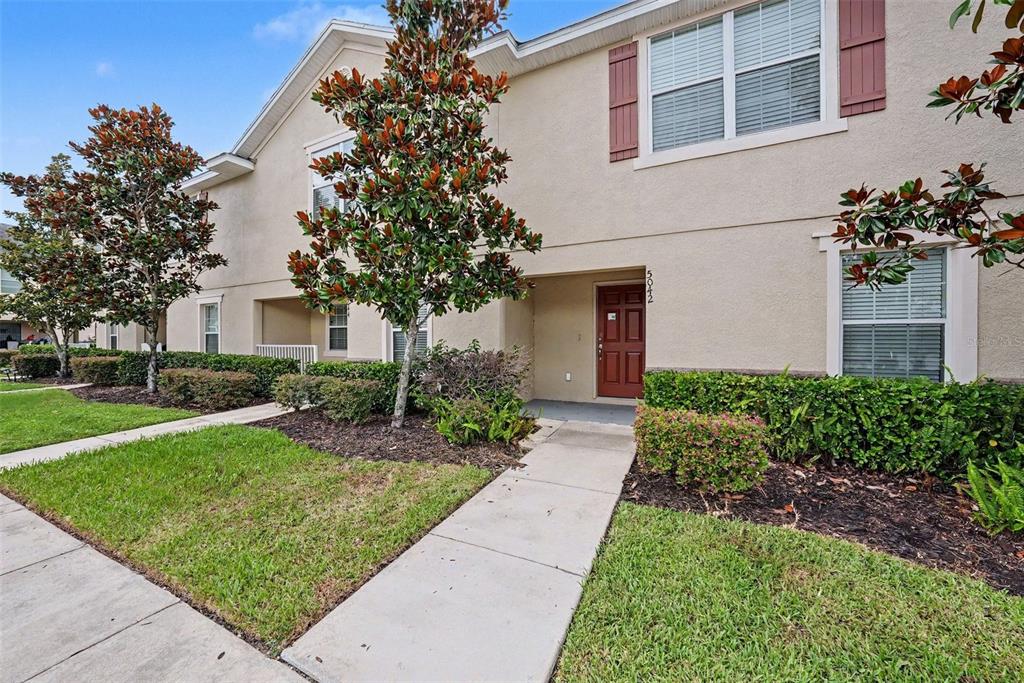 A two-story townhouse exterior with a red front door, tan stucco, and landscaped front yard — 5042 CHIPOTLE LANE, WESLEY CHAPEL, FL