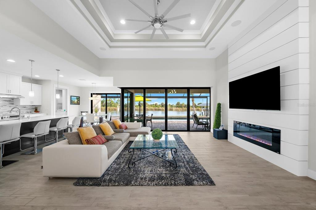 A modern open-concept living room features a white shiplap fireplace, tray ceiling, and sliding glass doors — 10572 RAINSVILLE STREET, PORT CHARLOTTE, FL