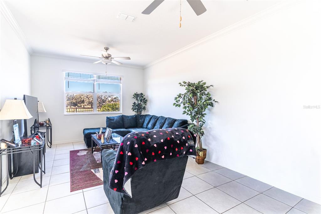 A living room features a dark sectional sofa, a patterned armchair, and tiled flooring — 7014 RIPPLE POND LOOP, ZEPHYRHILLS, FL