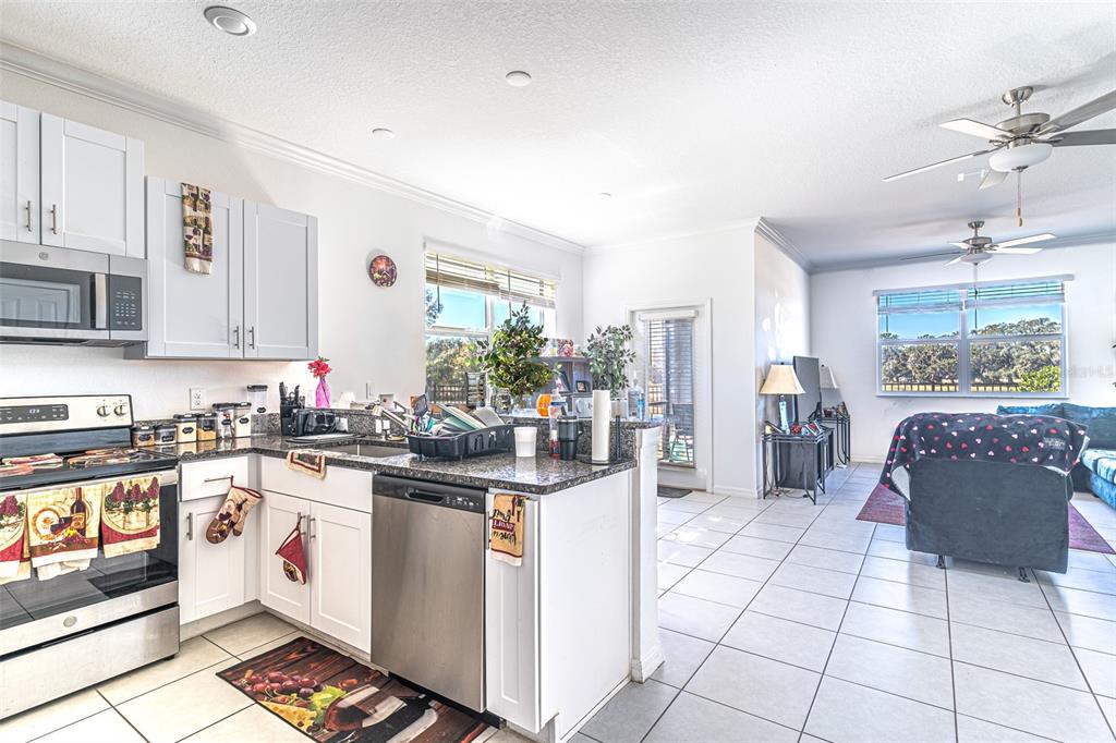 A kitchen with white cabinets and stainless steel appliances opens into a living room area — 7014 RIPPLE POND LOOP, ZEPHYRHILLS, FL