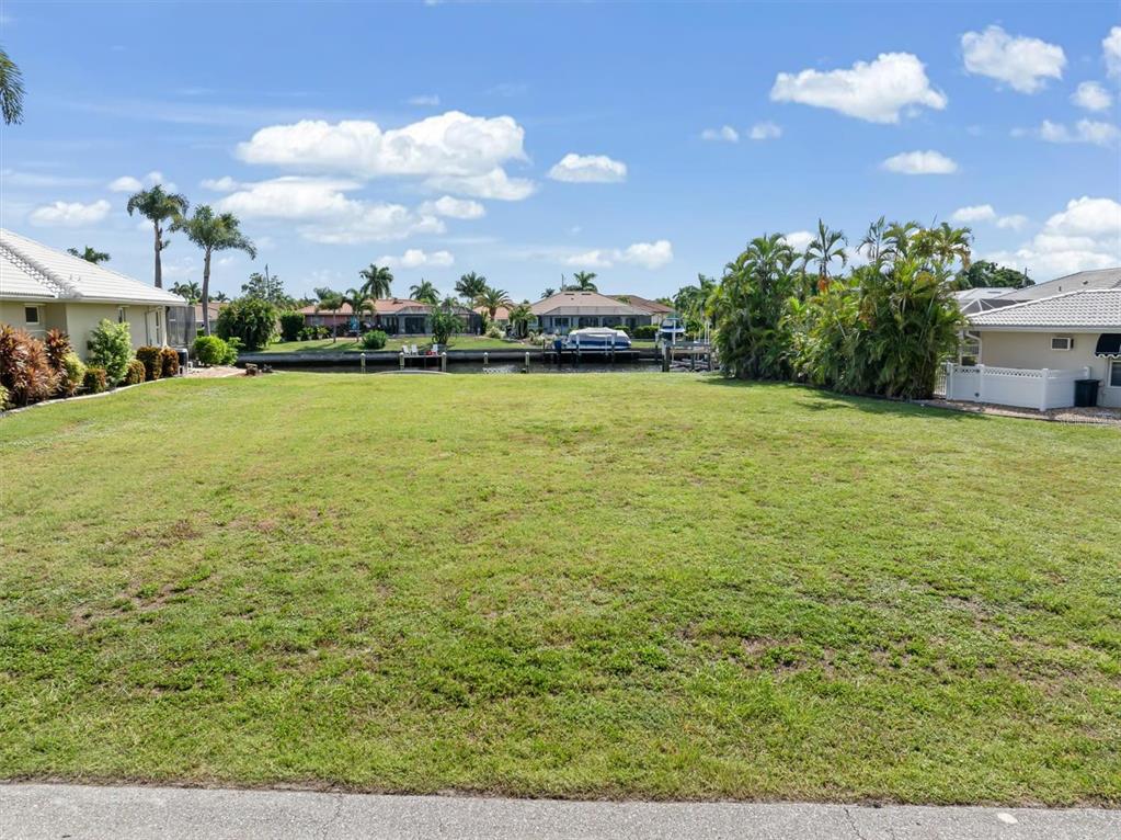 A grassy vacant waterfront lot sits between two residential houses under a blue sky — 834 SANTA MARGERITA LANE, PUNTA GORDA, FL