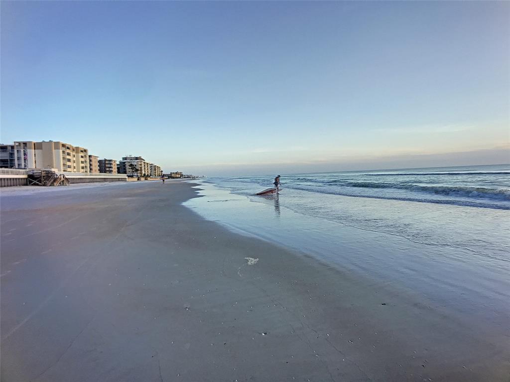 A person with a surfboard stands on a sandy beach near oceanfront apartment buildings — 4148 MICHIGAN AVENUE, NEW SMYRNA BEACH, FL