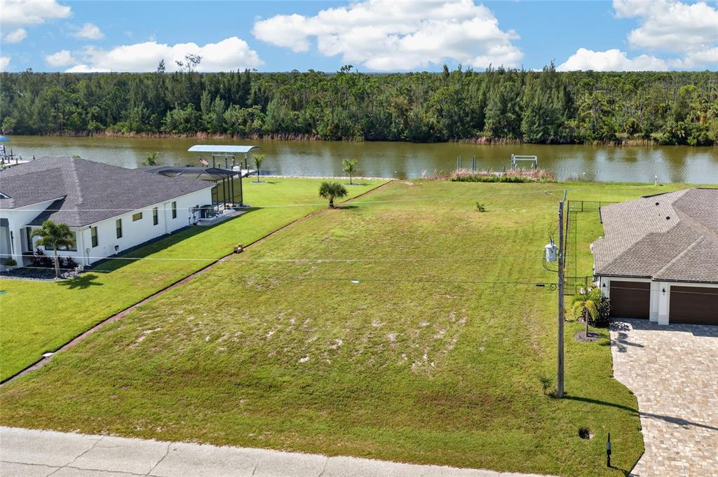 An empty residential lot sits between two houses along a canal with a treeline background — 16652 LIGGETT CIRCLE, PORT CHARLOTTE, FL